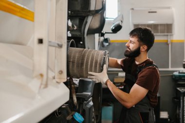Eldiven giyen oto tamircisi, yoğun bir garaj atölyesinde rutin bakım sırasında kirli hava filtresini değiştiriyor.