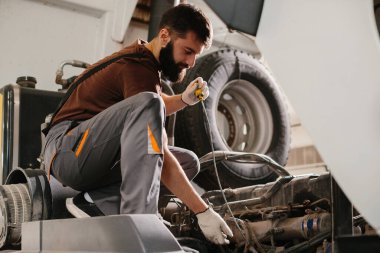 Tamirci bir kamyon motoru üzerinde çalışıyor, yağ seviyesini dipstick ile kontrol ediyor, tamirhanede bakım yapıyor.