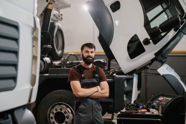 Bir kamyon servis merkezinde kolları çapraz duran gülümseyen bir tamircinin portresi ağır araç bakımında uzmanlık ve profesyonellik gösteriyor.