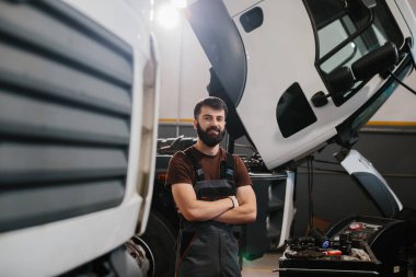 Meşgul bir kamyon tamirhanesinde çapraz silahlarla duran yetenekli bir tamircinin portresi otomotiv endüstrisinde uzmanlık ve profesyonellik sergiliyor.