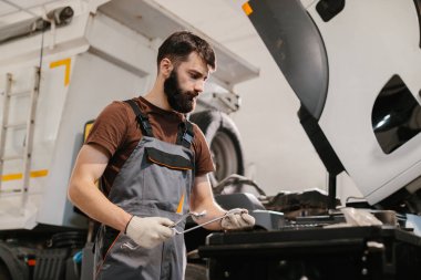 Tamirci yoğun bir garajda, aletler ve ekipmanlarla çevrili bir kamyon motoru üzerinde özenle çalışıyor.