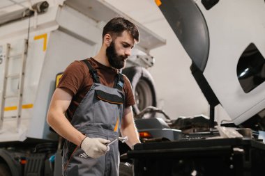 Otomobil tamircisi ingiliz anahtarı tutuyor, yoğun bir garaj atölyesinde bir kamyon motorunu özenle tamir ediyor, otomotiv tamirinde beceri ve uzmanlık gösteriyor.