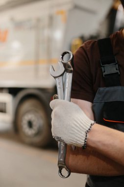 Tamirci eldiven takıyor ve İngiliz anahtarı tutuyor. İyi donanımlı bir garajda kamyon tamirine hazırlanıyor.