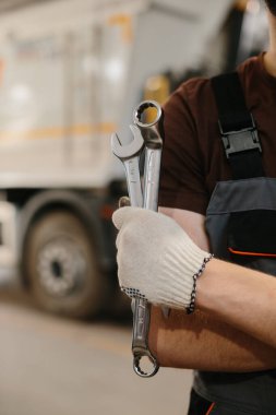 Bir atölyede araba tamircisi elinde iki büyük anahtar var. Kamyon tamirine hazır. Araç bakımında uzmanlık gösteriyor.