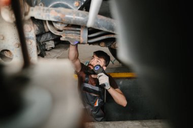 El feneri kullanan oto tamircisi, yoğun bir tamirhanede bir kamyonun altını dikkatle inceliyor, sorunları tanımlamaya odaklanıyor.