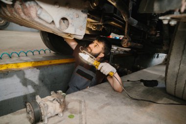 Tamir garajında bir kamyonun altını kontrol eden oto tamircisi, bileşenleri ve sorun giderme sorunlarını incelemek için çalışma ışığı kullanıyor.