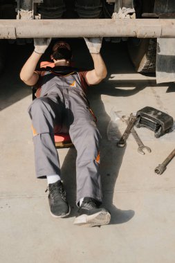 Otomobil tamircisi garajın zemininde yatıyor, anahtarı olan bir kamyonu tamir etmeye odaklanmış, kendini işine adamış.