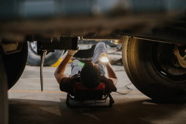 Bir kamyonun altında çalışan tamirci, bakımını yaparken aracın altını aydınlatmak için el feneri kullanıyor.