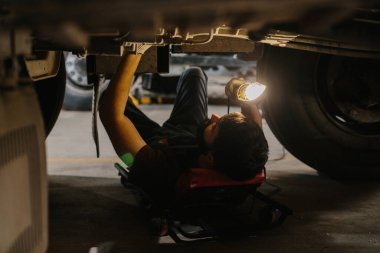 Bir kamyonun altında yatan tamirci, tamir alanını aydınlatmak için el feneri kullanırken şasiye ve sorun çıkarma sorunlarına odaklanıyor.