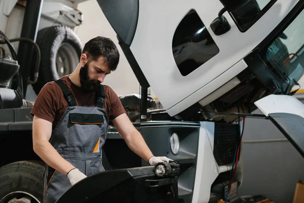 Otomobil tamircisi garajda bir kamyon motoru üzerinde çalışıyor. Aracı incelemek ve onarmak için el feneri ve aletler kullanıyor.