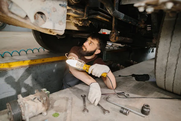 Deneyimli bir tamirci, büyük bir kamyonun altında çalışır. Alet ve lamba kullanarak aracın bagajını kontrol eder ve tamir eder.