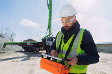 Çelik yelek ve kask giyen inşaat işçisi beton üretim tesisinde uzaktan kumandalı beton pompa kamyonu kullanıyor.