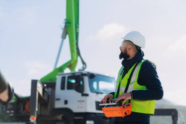 Güvenlik kaskı ve yelek giyen inşaat işçisi beton üretim tesisinde uzaktan kumandalı beton pompa kamyonu kullanıyor.