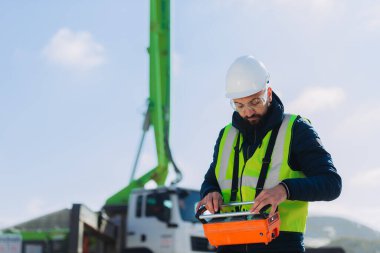 İnşaat işçisi inşaat alanında uzaktan kumandayla beton pompa kamyonu kullanıyor, güvenlik kaskı ve yelek giyiyor.