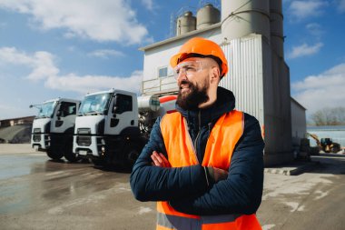 Güvenlik kaskı ve yelek giyen kendine güvenen inşaat işçisi beton üretim tesisinde çapraz kolları olan, arka planda park etmiş beton karıştırıcı kamyonlar.