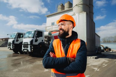 Güvenlik kaskı ve yelek giyen kendine güvenen bir yönetici beton üretim tesisinde çapraz kollarla duruyor, kamyonları ve çimento karıştırıcıları denetliyor.