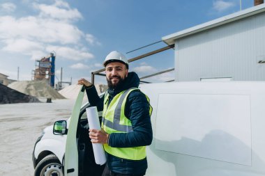 Elinde somut bir üretim fabrikasının planlarıyla şirketin minibüsünden çıkan gülümseyen bir mühendis.