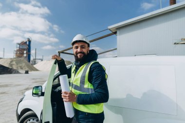 Minibüsünün yanında beton üretim tesisinde duran gülümseyen mühendis elinde planlar tutuyor.