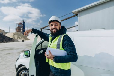 Beton üretim tesisinde minibüsünün yanında duran kendine güvenen mühendis, endüstriyel kalkınma ve inşaatı sembolize ediyor.