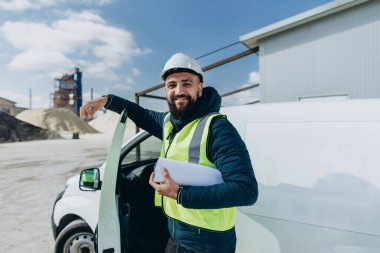 Güneşli bir günde beton üretim tesisinde minibüsünün yanında duran gülümseyen mühendis elinde planlar tutuyordu.