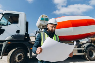 İnşaat mühendisi güvenlik kaskı ve yelek giyiyor inşaat alanındaki beton karıştırıcı kamyonun yanındaki planı inceliyor.