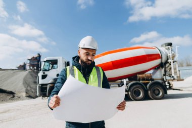 Güvenlik kaskı ve yelek giyen inşaat mühendisi beton üretim tesisinin planını inceliyor.