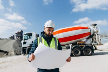 Güvenlik kaskı ve yelek giyen inşaat mühendisi arka planda çimento karıştırıcı kamyonu olan beton üretim fabrikasının planlarını inceliyor.