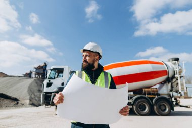 Güvenlik kaskı ve yelek giyen inşaat mühendisi, beton karıştırıcı kamyonu ve çöp kamyonu olan bir fabrikada beton üretimini denetliyor.