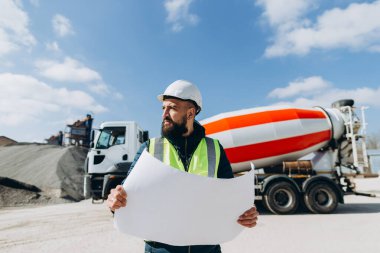 Çelik yelek ve miğfer giyen inşaat mühendisi, beton üretim tesisinde beton karıştırma kamyonunu denetlerken elinde taslak tutuyor.