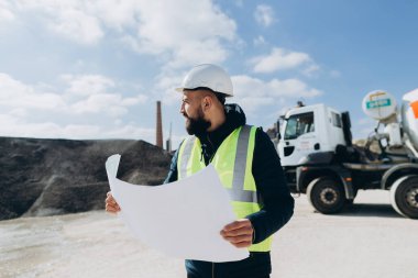 Güvenlik yeleği ve kask giyen inşaat mühendisi, bir beton üretim fabrikasının planlarını inceliyor ve proje uyumunu ve kalitesini sağlıyordu.