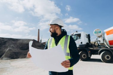 Güvenlik kaskı ve yelek giyen bir müfettiş arka planda çimento karıştırıcı kamyonu olan beton üretim tesisini denetleyen bir plan tutuyor.