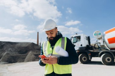 Emniyet yeleği ve şapka giyen inşaat mühendisi, kolunun altında planları olan beton üretim tesisinde akıllı telefon kullanıyor.
