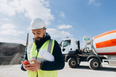 İnşaat mühendisi planlar tutuyor ve arka planda çimento kamyonu olan bir beton üretim tesisinde akıllı telefon kullanıyor.