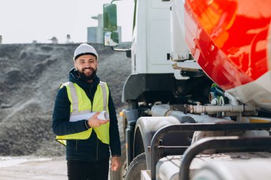Güvenlik kaskı takan inşaat mühendisi ve yüksek görünürlüklü yelek elinde beton fabrikasının beton üretimini denetleyen planlar var.