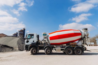 Mavi gökyüzünün altındaki beton üretim tesisinde çakıl yığınının yanına park edilmiş kırmızı ve beyaz beton karıştırıcı kamyon.