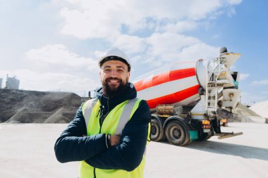 Beton üretim tesisinde çimento kamyonunun önünde kollarını kavuşturmuş gülümseyen bir mühendisin portresi.