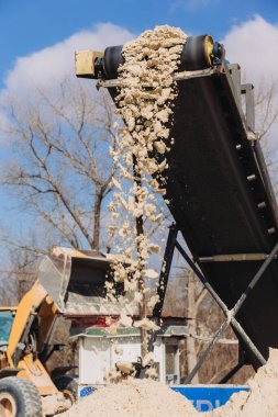 Taşıyıcı bant, beton üretim tesisinde kum yığınına dökülüyor. Arka planda buldozer ve mavi gökyüzü var.
