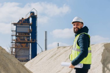 Mühendis, güneşli bir günde beton üretim fabrikasının önünde elinde bir plan tutarken gülümsüyor.