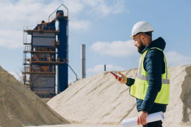 İnşaat mühendisi akıllı telefon kullanıyor ve bir beton üretim fabrikasının planlarını tutuyor, endüstriyel operasyonları yönetiyor ve denetliyor.