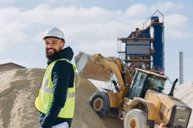 İnşaat müfettişi güneşli bir günde kazıcı yakınında gülümsüyor ve beton üretim tesisinde kum yığını var.