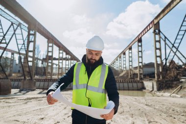 Güvenlik yeleği ve kask giyen inşaat mühendisi beton üretim tesisinin planlarını inceliyor.