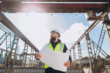 Güvenlik kaskı ve yeleği takan gülümseyen mühendis, beton üretim tesisinin planlarını tutuyor, inşaat alanını inceliyor.