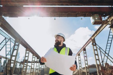 İnşaat mühendisi elinde parlak güneş ışığı altında beton üretim tesisini denetleyen bir proje tutuyor.