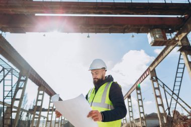 Güvenlik kaskı ve yelek giyen inşaat mühendisi güneşli bir günde beton üretim tesisinin planlarını inceliyor.
