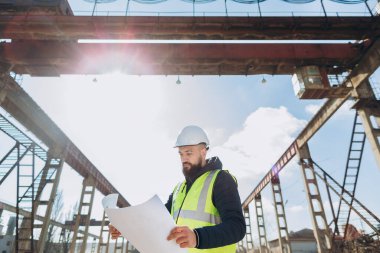 Kask ve güvenlik yeleği giyen mühendis, güneşli bir günde beton üretim fabrikasının planlarını inceliyor.