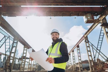 Güvenlik kaskı ve yelek giyen gülümseyen mühendis beton üretim tesisinin planlarını elinde tutuyor.