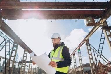 Güvenlik yeleği ve miğfer giyen gülümseyen mühendis beton üretim tesisinde vinç altında plan inceliyor.