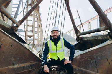 Gülen inşaat işçisi çelik yelek ve şapka takıyor beton üretim tesisinde ağır makinelerin üzerinde oturuyor, endüstriyel çevreyi gösteriyor.