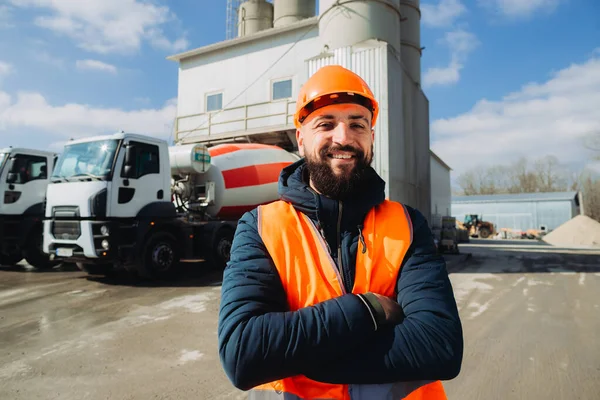 Beton üretim tesisi önünde çimento karıştırıcı kamyonları ve siloları ile çapraz gülümseyen kendinden emin inşaat işçisi