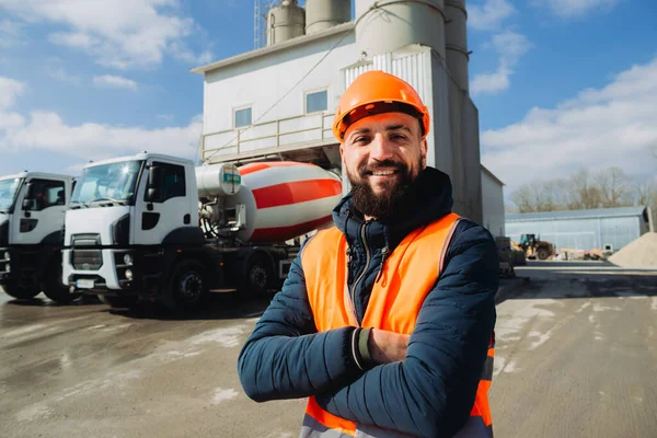 Çimento kamyonları ve beton üretim tesisi önünde çapraz kollarla gülümseyen kendinden emin inşaat işçisi